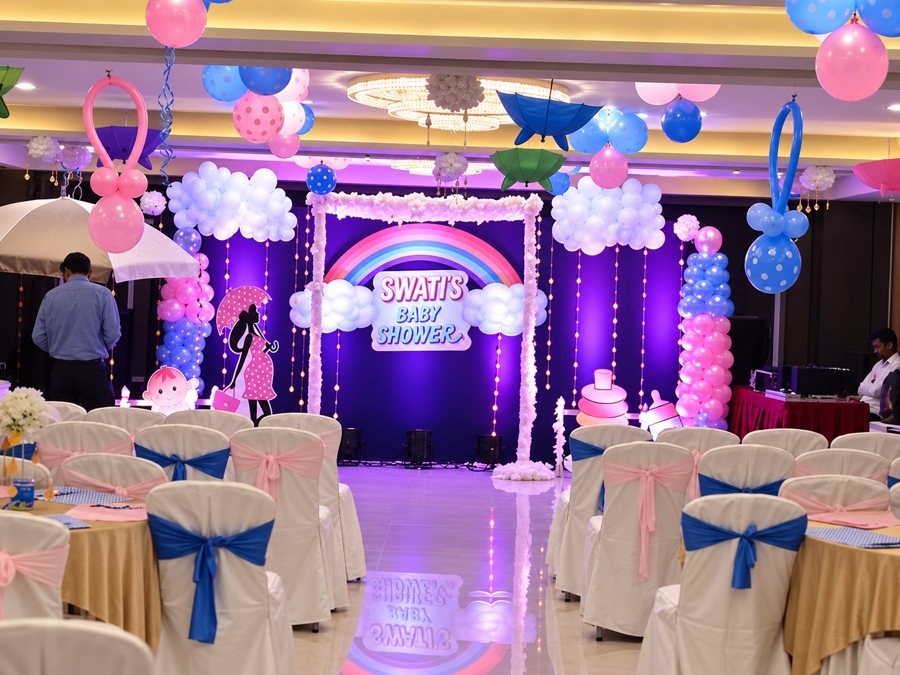 Baby shower hall decor with a rainbow stage backdrop, balloon clouds, balloon pillars and pink and blue chair sashes